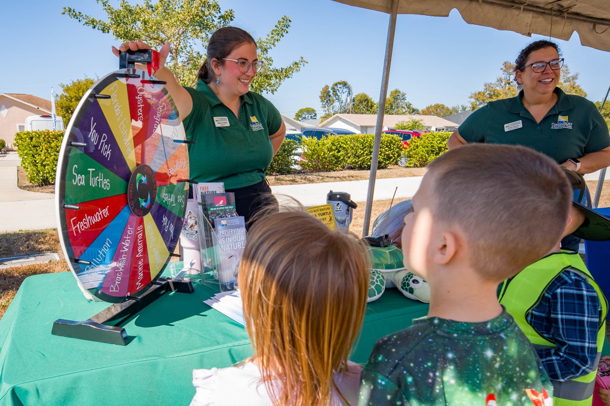 Two smiling staff members in green shirts run a prize wheel with topics like sea turtles, oysters and seagrass for children at a Waterways Festival booth.